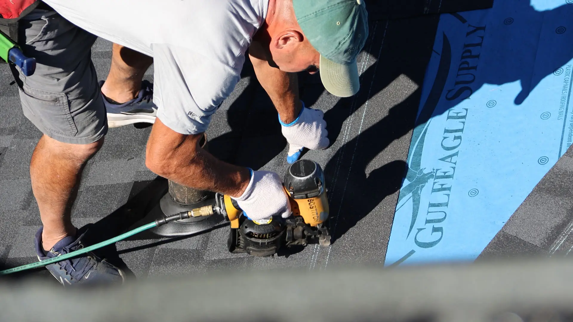 Close-up of roofer using nail gun to fasten shingles near roof vent.
