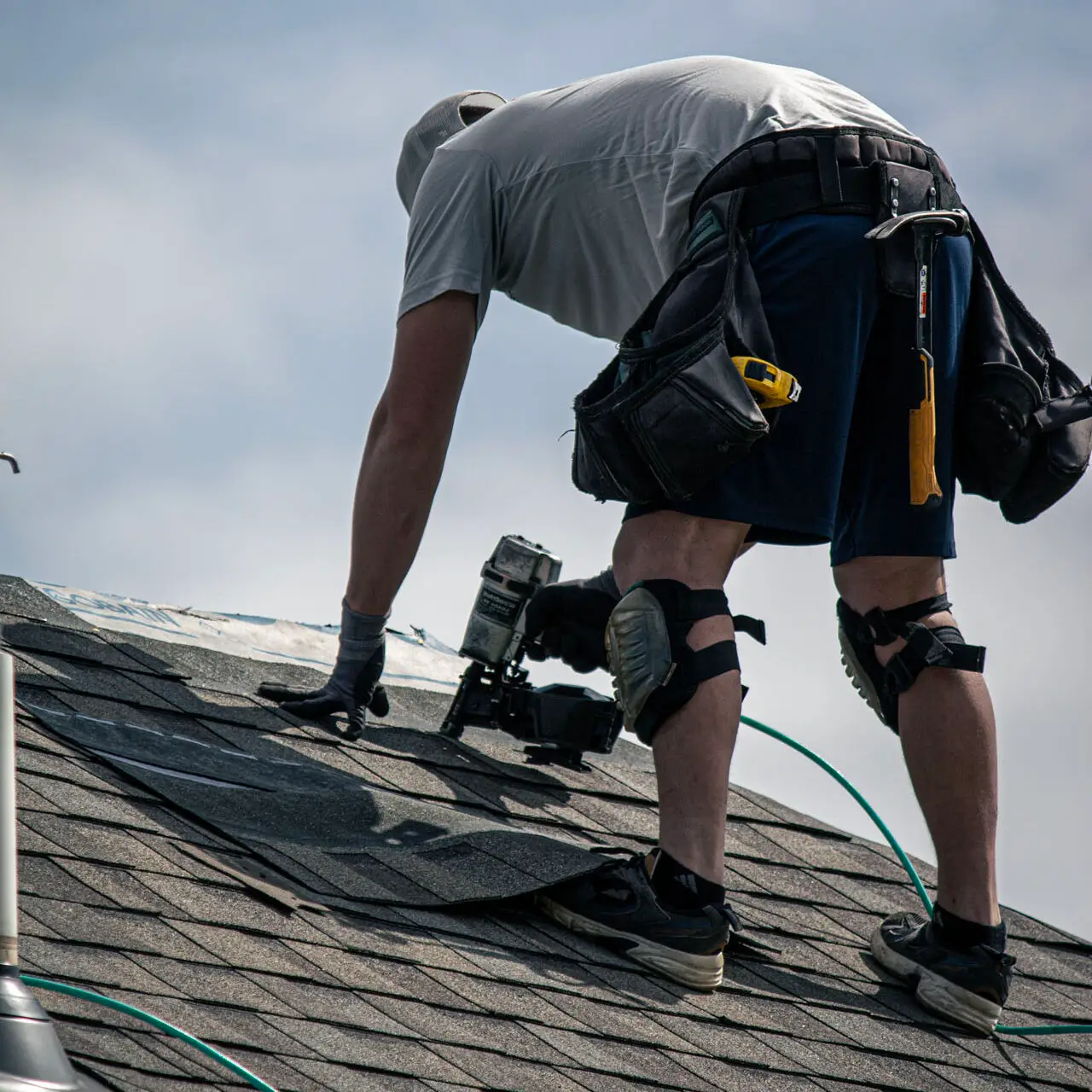 Roofer using a nail gun to secure asphalt shingles near the roof ridge on a residential home.