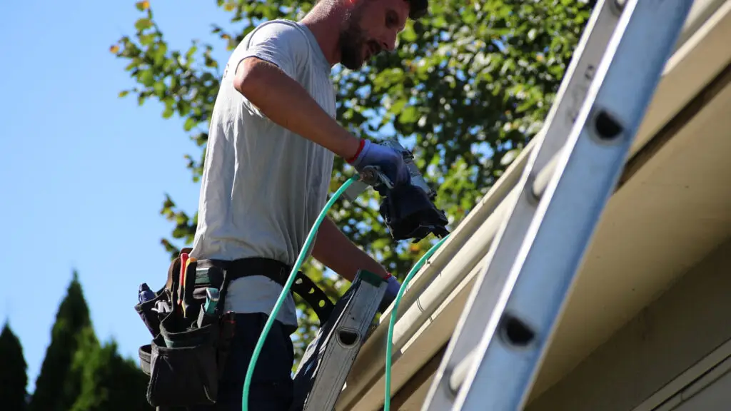 Roofer installing asphalt shingles with a nail gun along roof edge in Burton, MI.