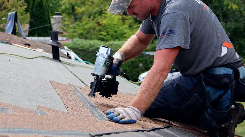 Roofer installing asphalt shingles with nail gun on residential roof in Deerfield, MI.