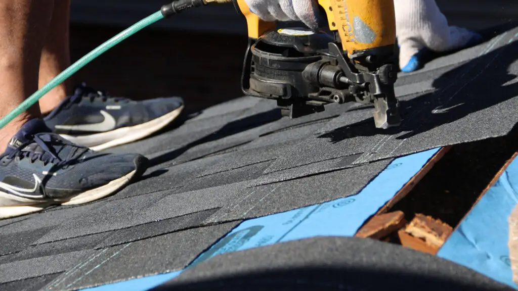 Roofer secures asphalt shingles with nail gun near roof ridge in Huntington Woods, MI.