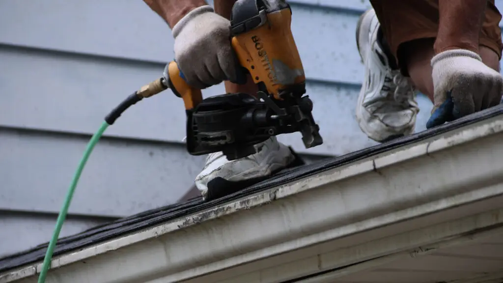Roofer securing asphalt shingles with a nail gun at the roof eave in Flint, MI.