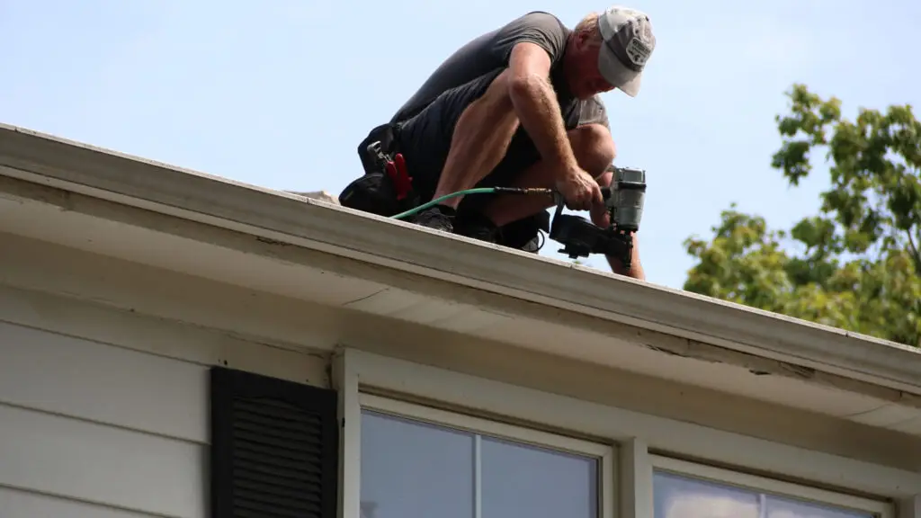 Roofer securing asphalt shingles with nail gun along roof eave in Farmington Hills, MI.