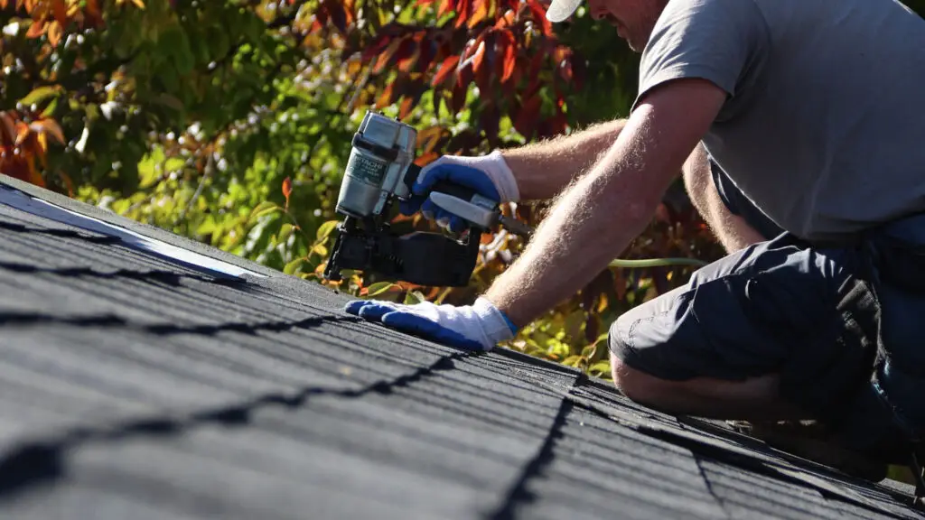 Roofer using nail gun to secure asphalt shingles by roofs edge in Waterford, MI.