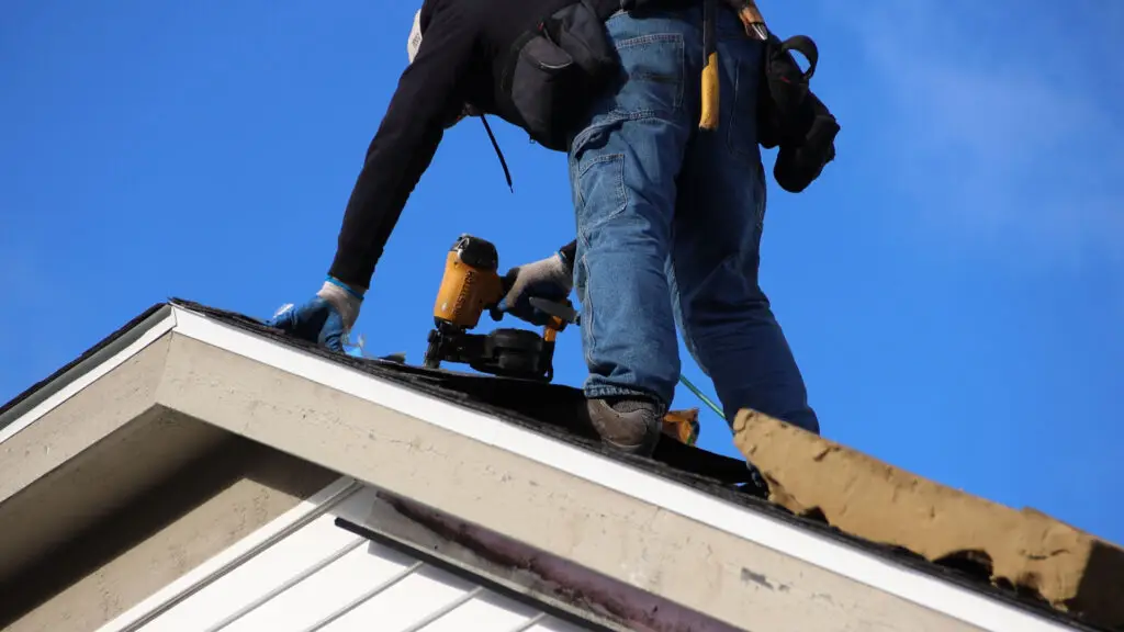 Roofer using nail gun to secure asphalt shingles near roof ridge in Ferndale, MI.