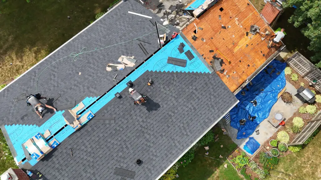 Roofing crew installing new shingles on the main roof while another crew member strips old shingles from the lower roof in Grand Blanc, MI.